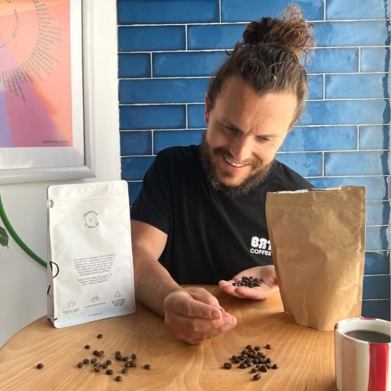 Tom Saxon of Batch Coffee Club looking at a handful of roasted coffee beans beside two coffee bags and a cup of black coffee